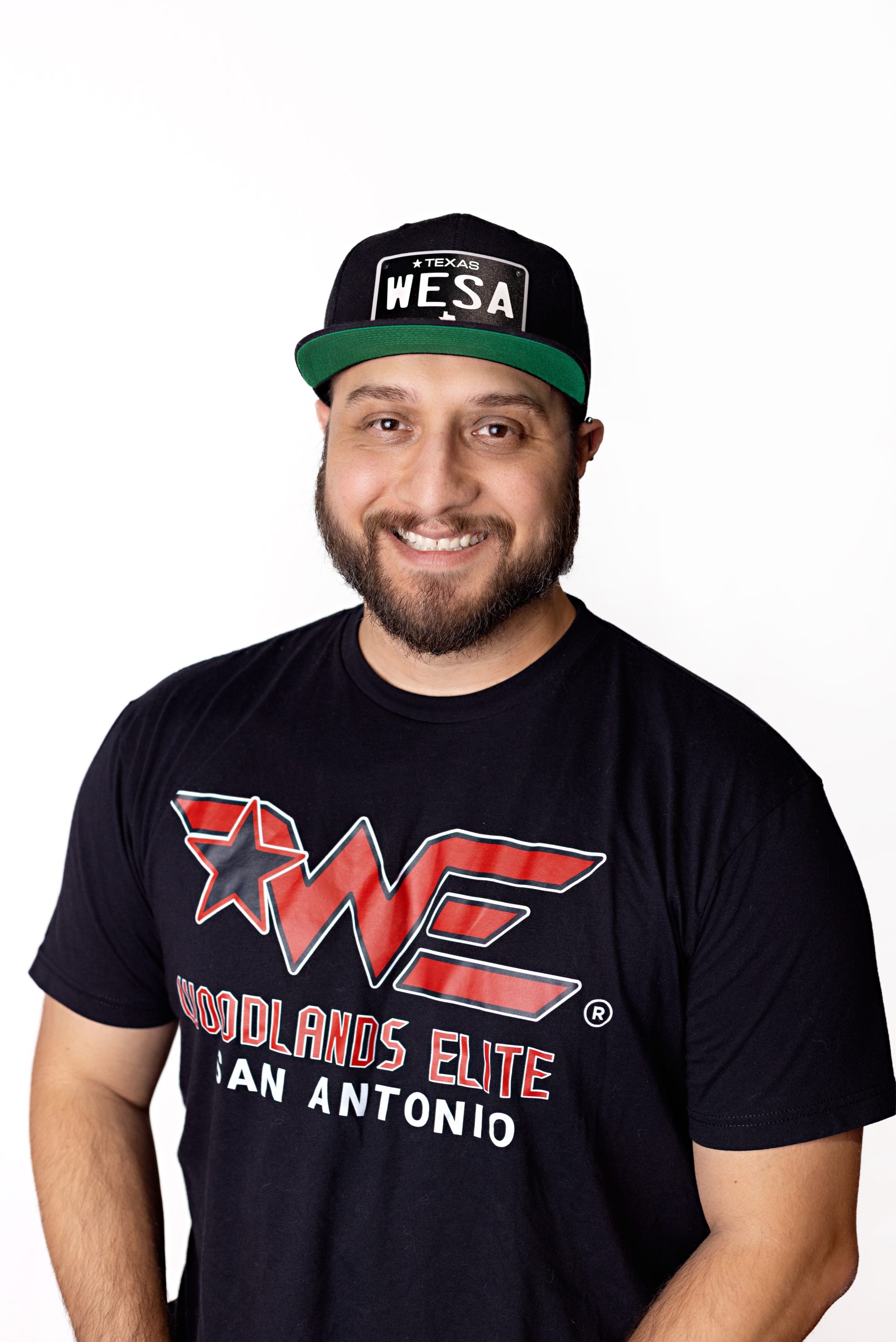 Smiling man wearing a black cap with a white WESA logo and a black T‑shirt with a red 'Woodlands Elite San Antonio' graphic.